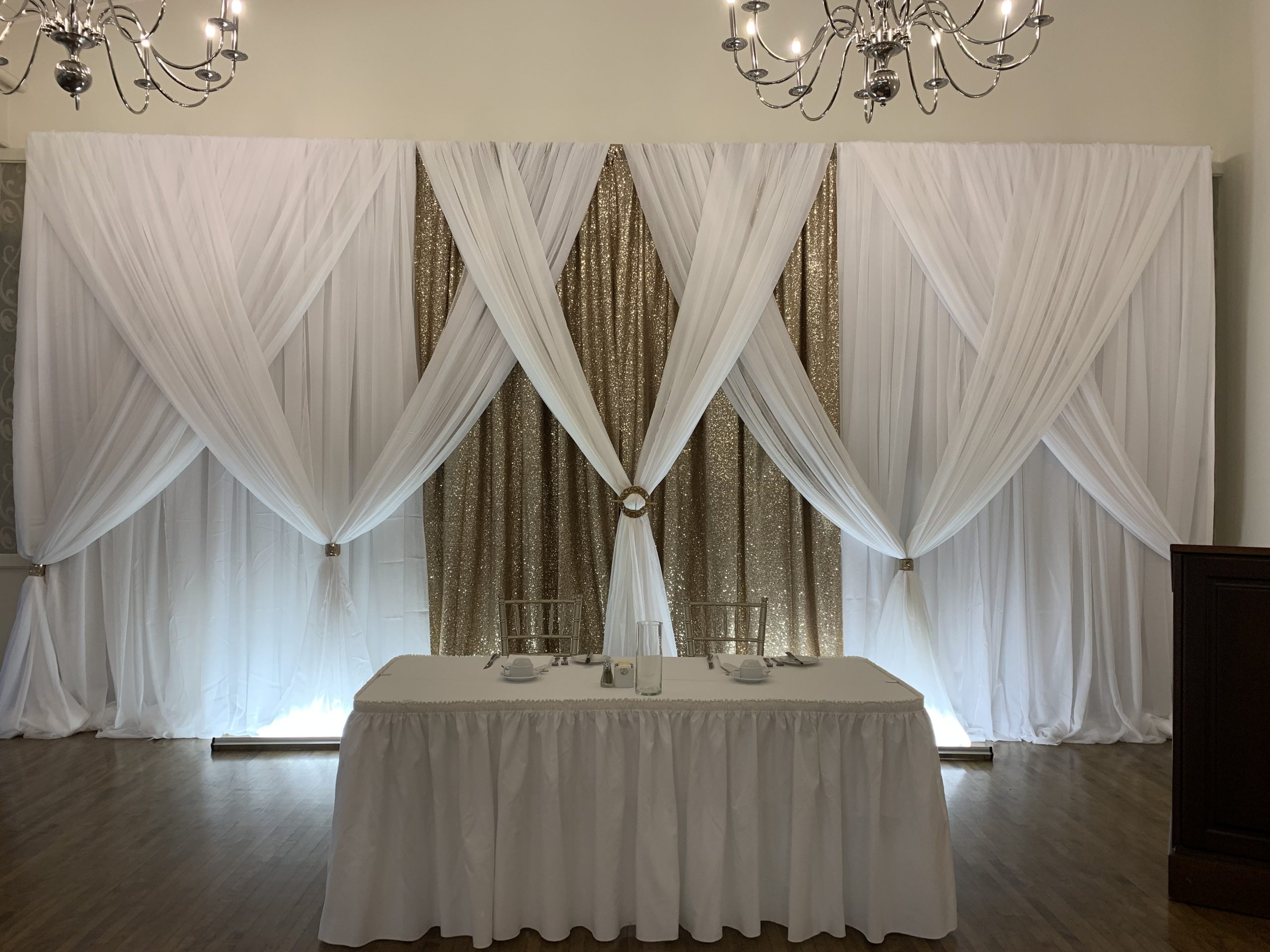 White and gold backdrop and table rentals used as a head table during a wedding reception.